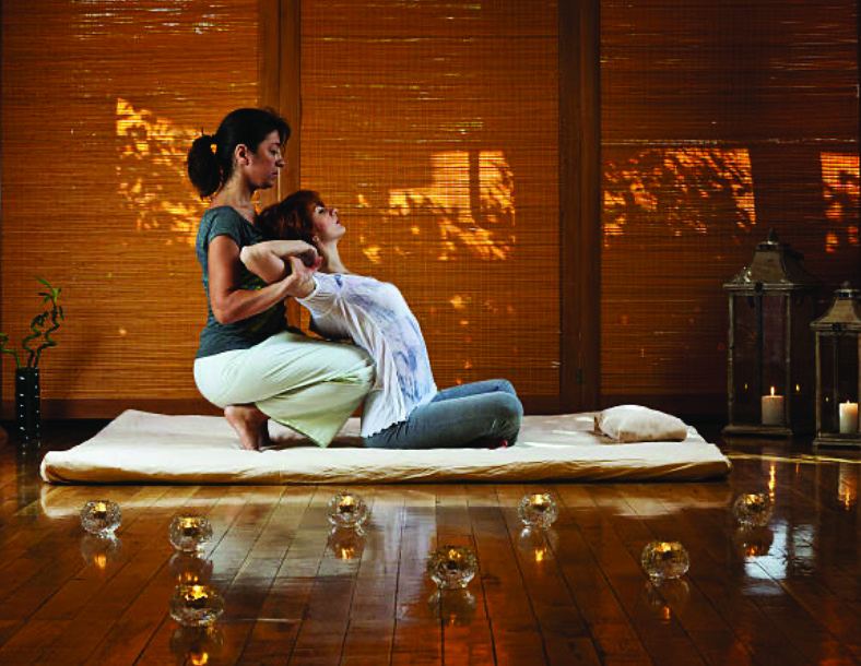 A therapist performing assisted Thai yoga stretching on a floor mat in warm candlelight